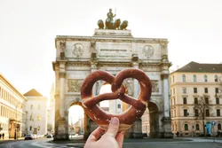 German pretzel in the hand against the backdrop of the Victory Gate triumphal arch Siegestor in Munich. Germany