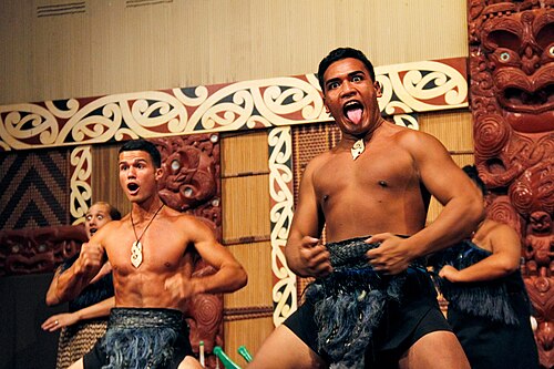 Māori dancers performing the Haka (traditional war cry). The dancers are wearing the traditional Maori necklaces known as the Hei matau and as well their traditoinal garb; fists are clenched and tongues are out.